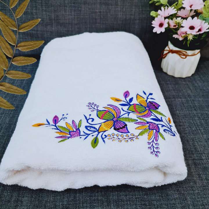 Floral embroidered bath towel folded with a golden leaf decor on a dark background.